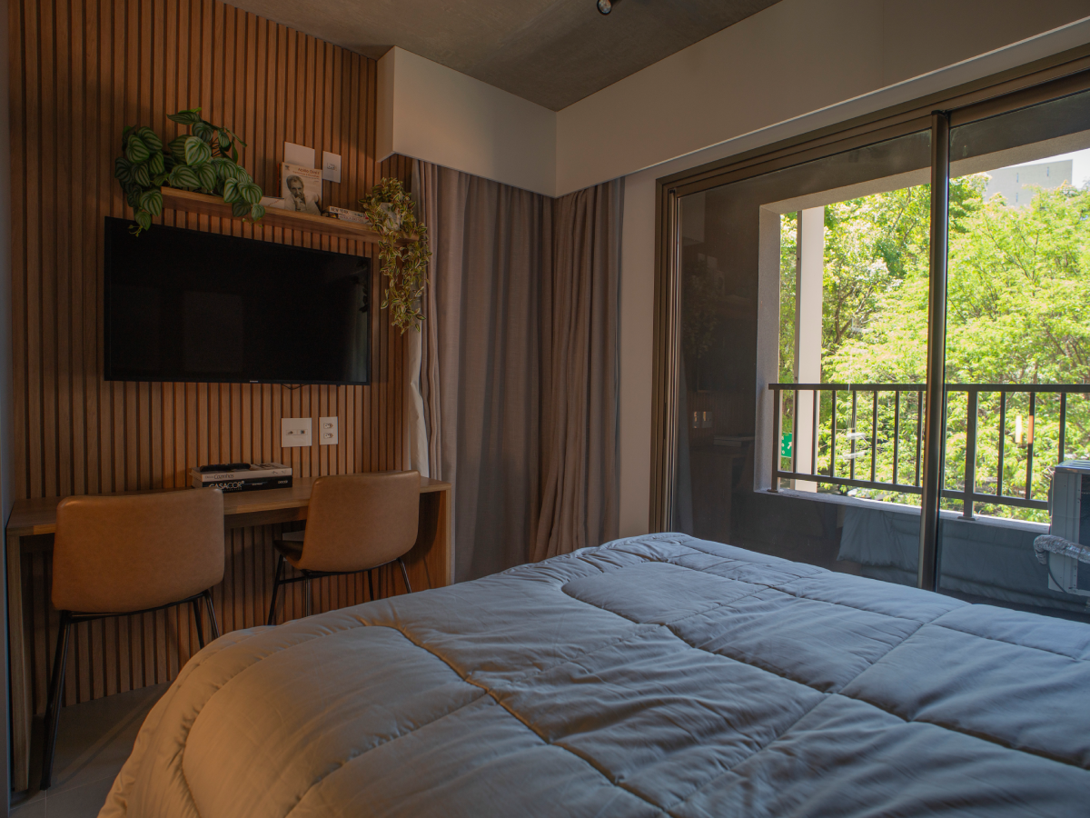 Quarto aconchegante com cama em primeiro plano, painel de madeira ripada com TV e bancada de trabalho com duas cadeiras, além de uma ampla porta de vidro que dá acesso à varanda com vista para árvores.