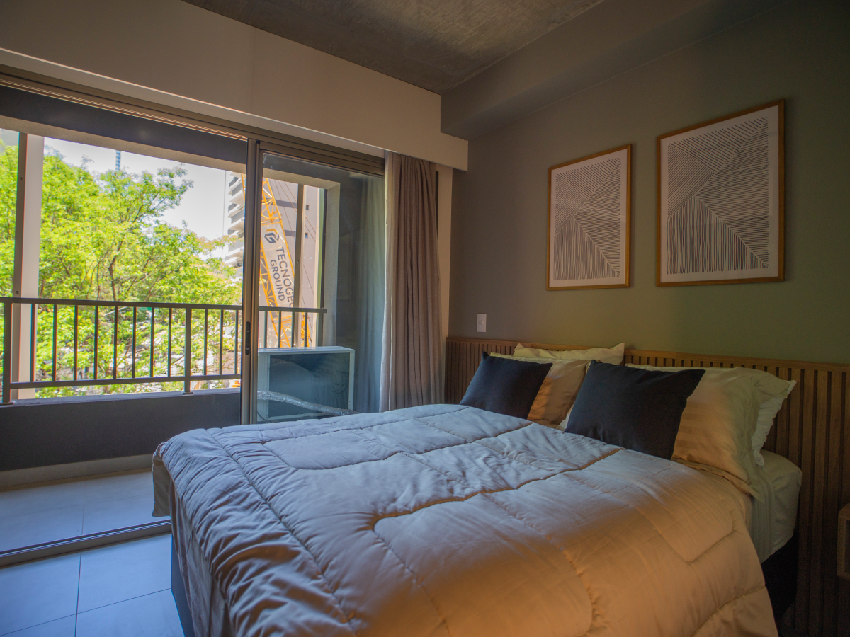 Quarto moderno com cama de casal arrumada, cabeceira de madeira ripada e quadros geométricos na parede cinza. Ao fundo, ampla porta de vidro dá acesso à varanda com vista para árvores verdes.