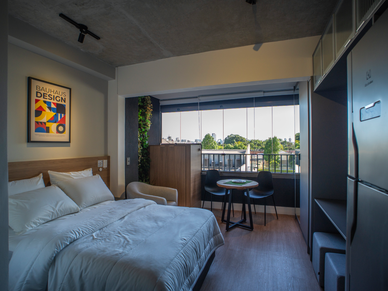 Quarto de apartamento studio moderno com cama de casal, quadro da Bauhaus na parede, teto de concreto aparente e varanda integrada com mesa pequena.