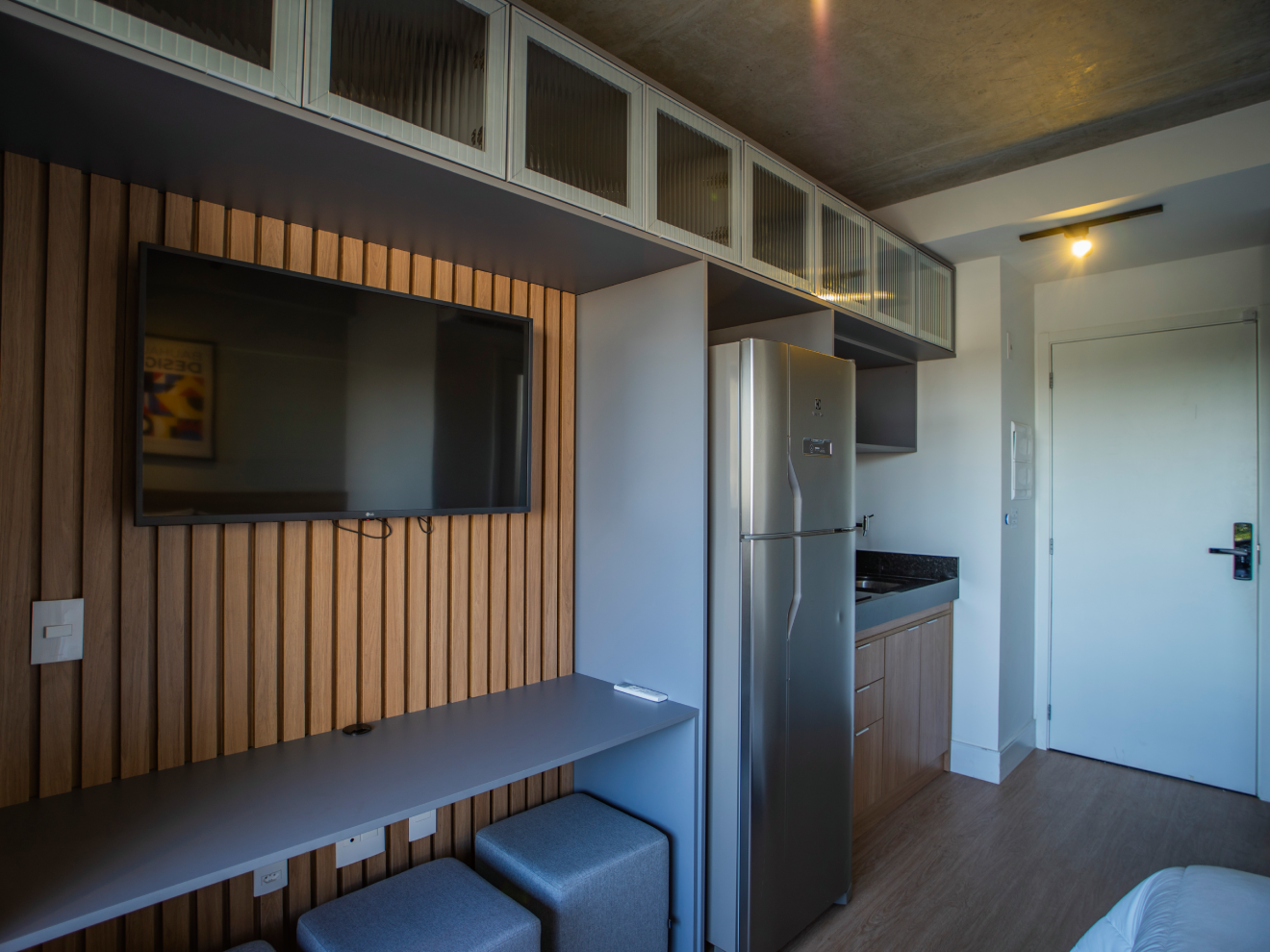 Interior de apartamento compacto com painel de TV em madeira ripada, bancada cinza com pufes e cozinha integrada com geladeira em inox.