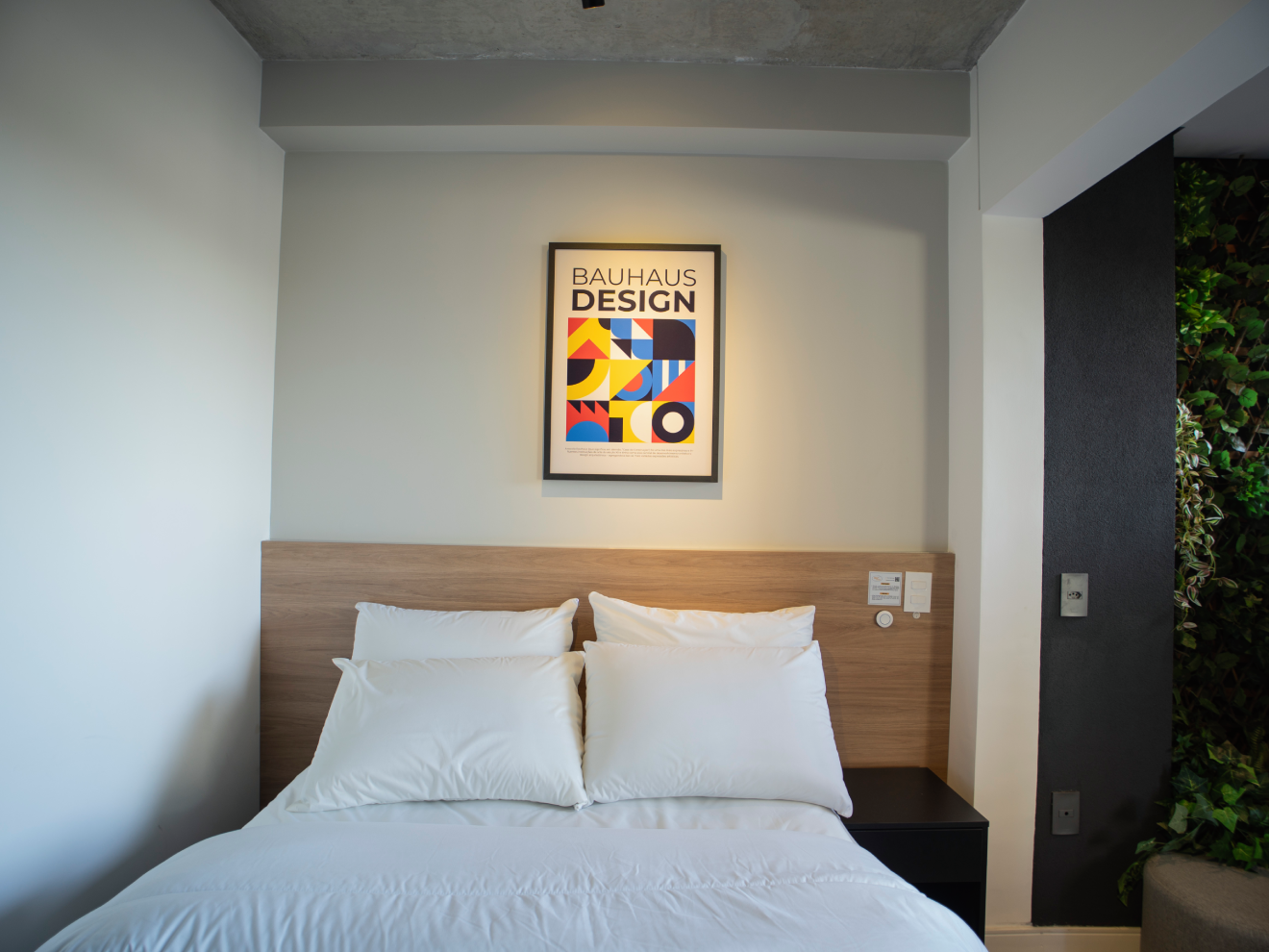 Cama de casal com roupa de cama branca, cabeceira de madeira e quadro "Bauhaus Design" em destaque na parede clara.